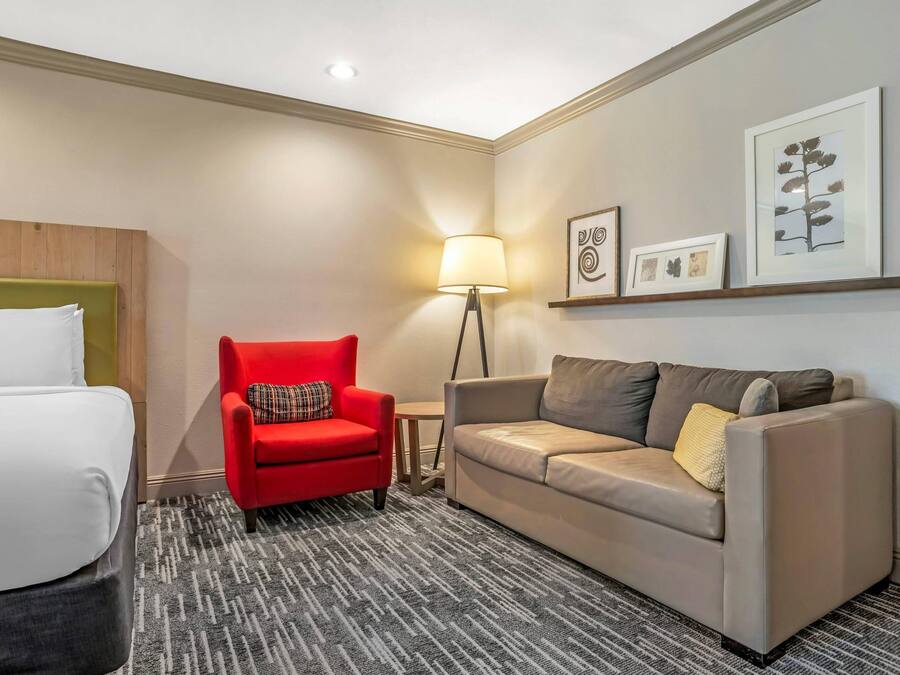 A hotel room with a bed, a red armchair, a beige sofa, a floor lamp, and framed artwork on the wall.