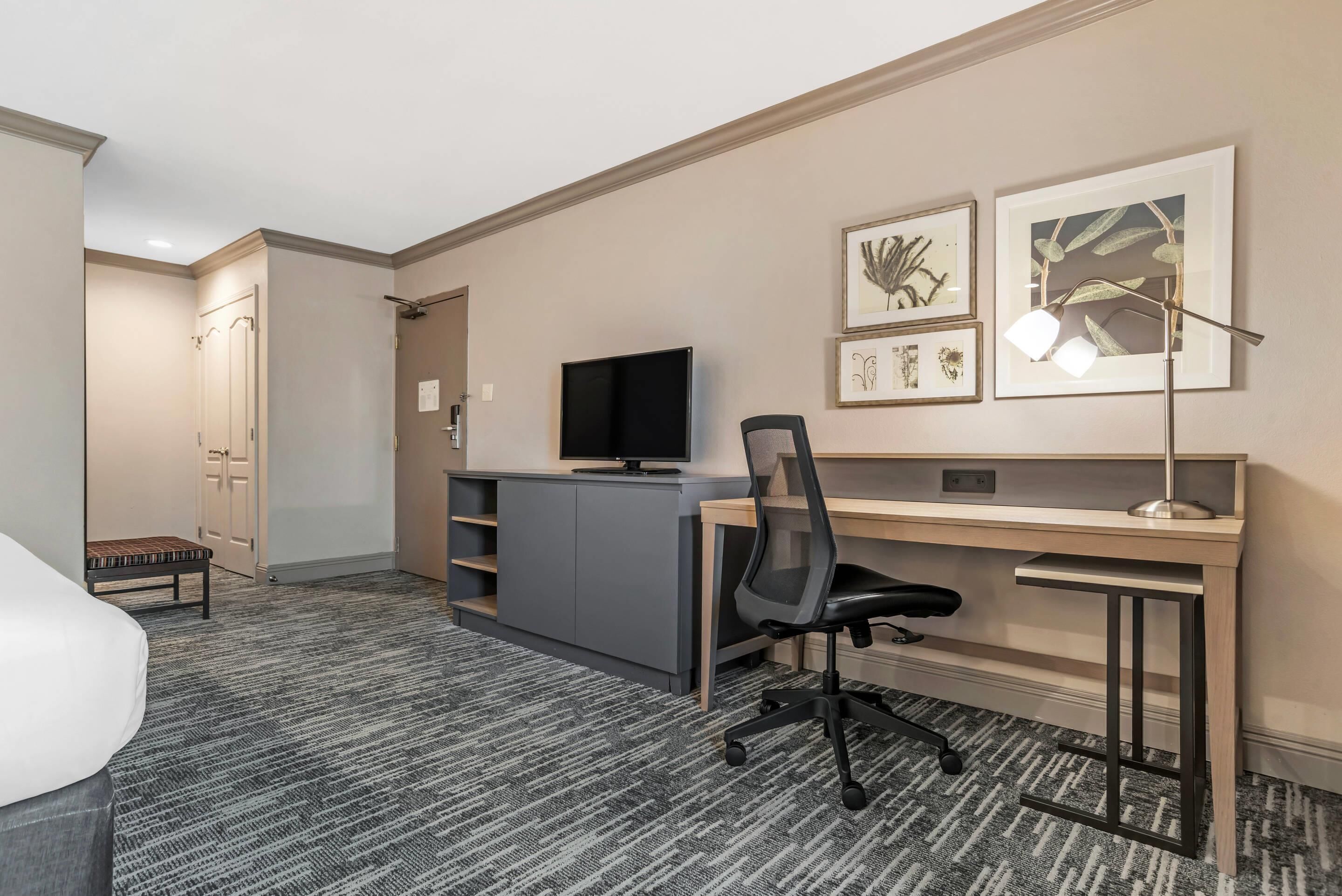 A hotel room with a bed, a desk with a computer, a chair, a TV, and framed artwork on the wall.