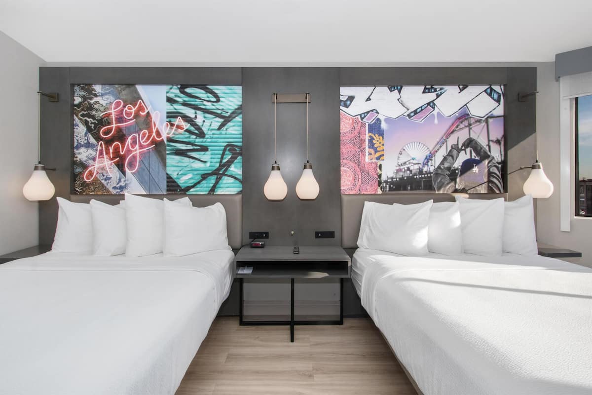 A modern hotel room with two beds, a small desk, and artwork on the walls.