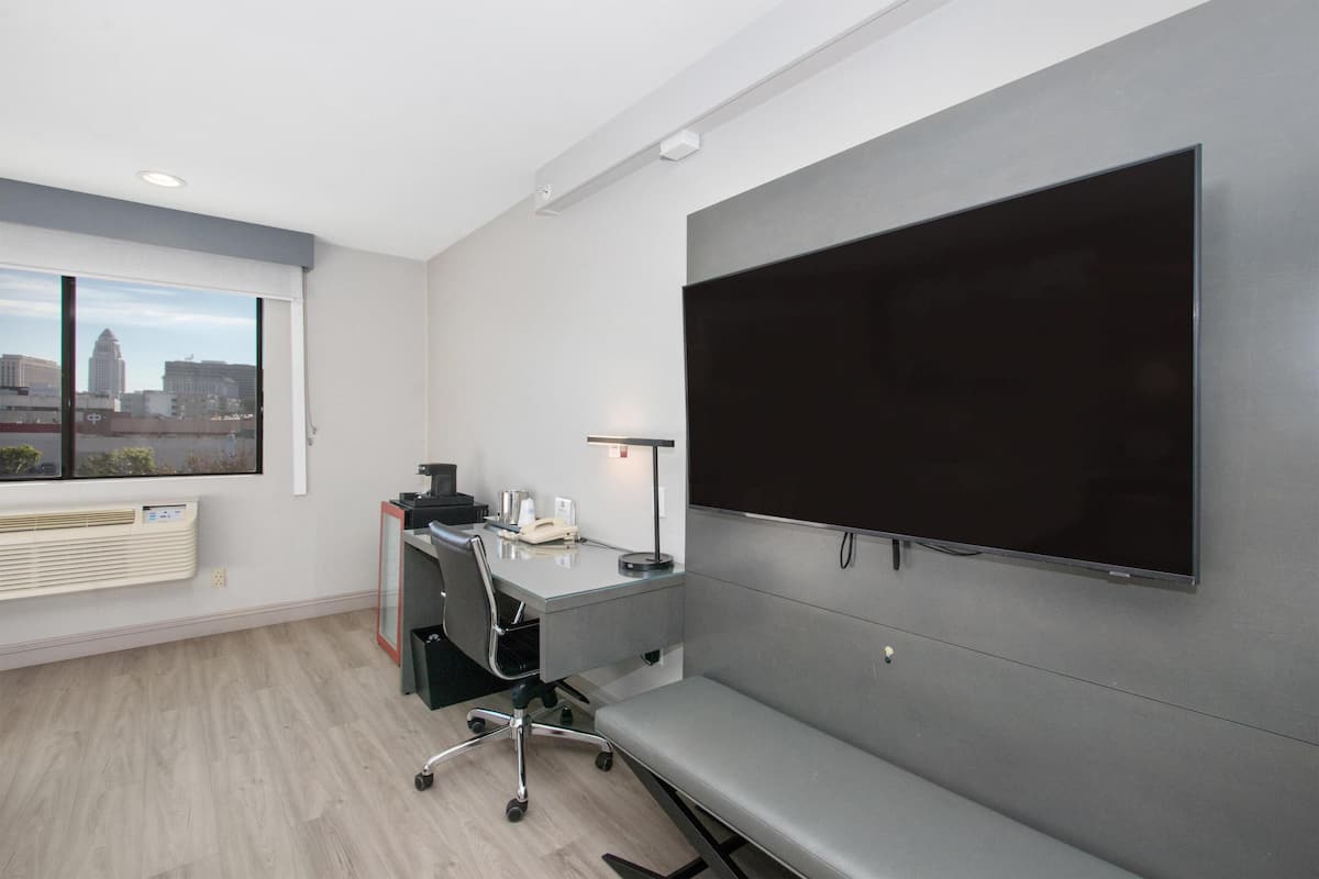 A modern office space with a large flat-screen TV mounted on the wall, a desk with a chair, a small table with a lamp, and a window with a view of the city skyline.