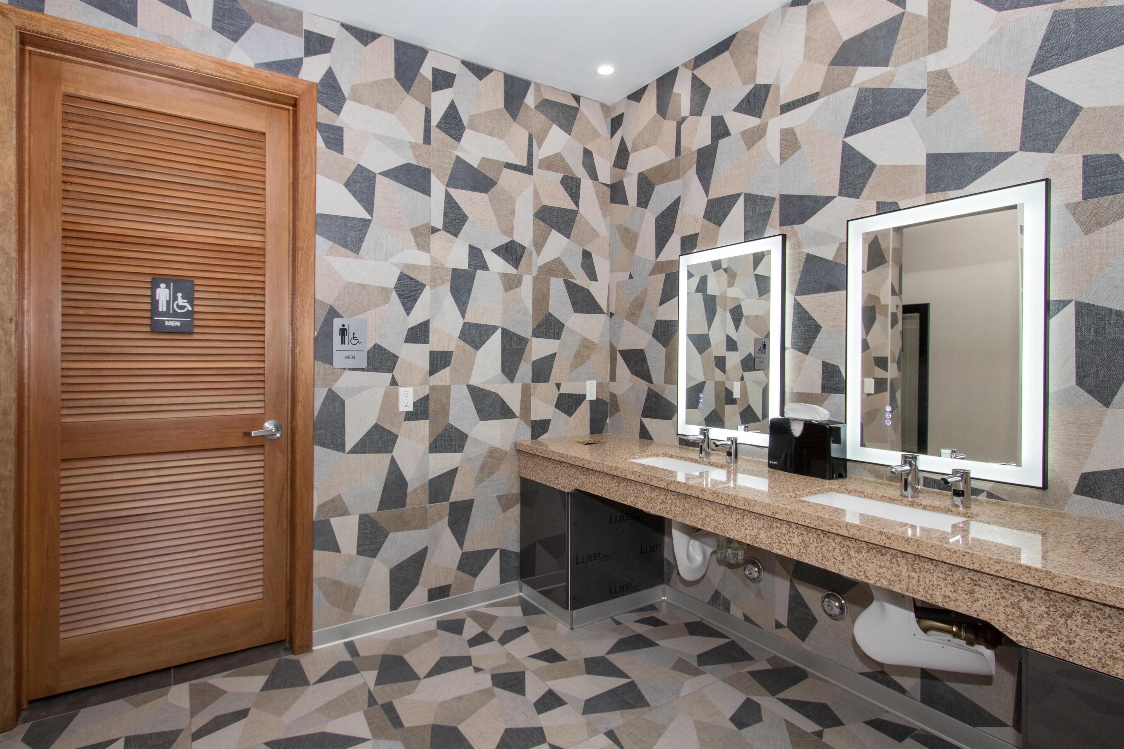 Une salle de bain moderne avec des murs aux motifs géométriques, une porte en bois et un meuble-lavabo doté de deux miroirs.