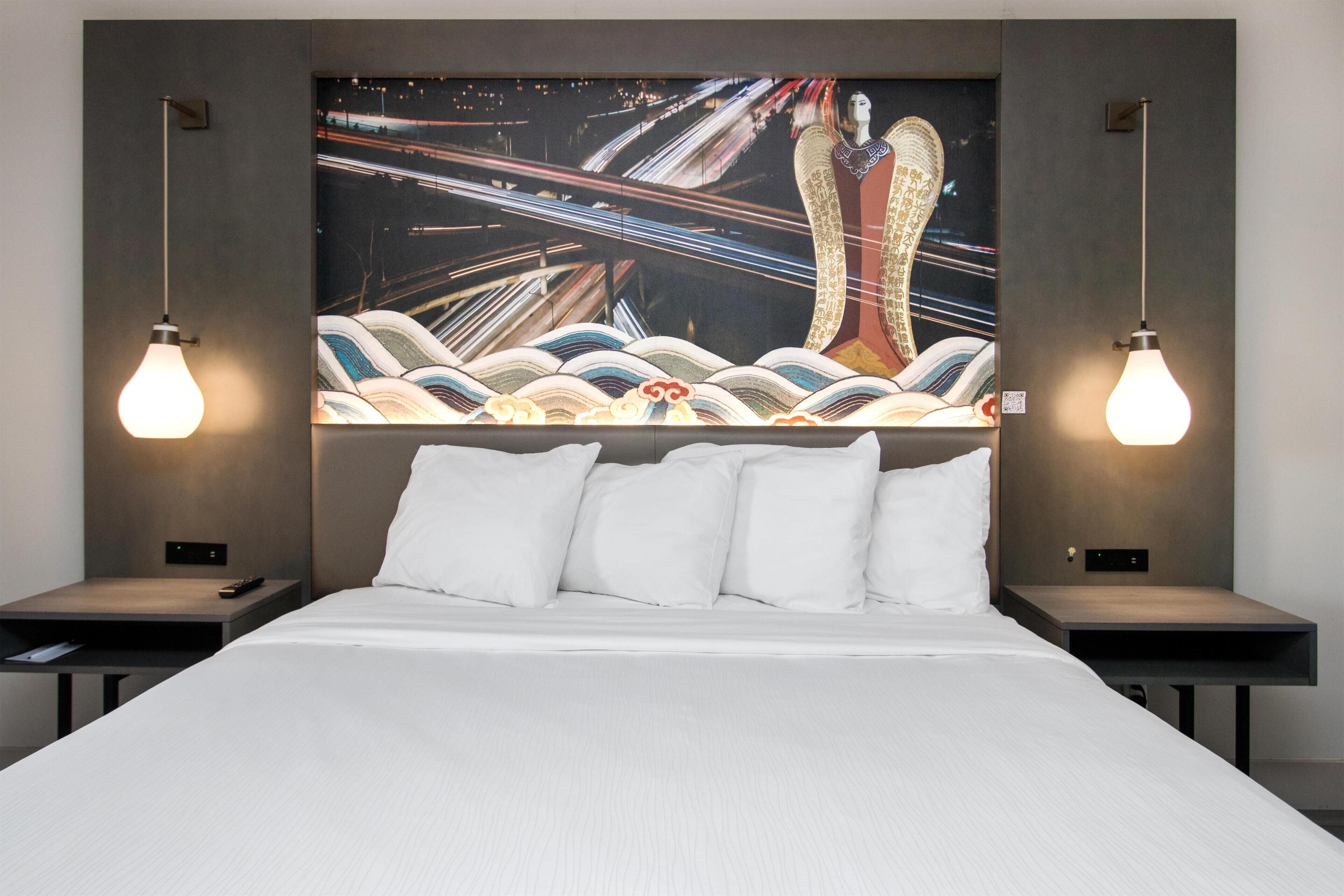 Una habitación de hotel moderna con una cama grande, ropa de cama blanca, dos mesitas de noche y un cuadro colgado en la pared con una vista urbana.