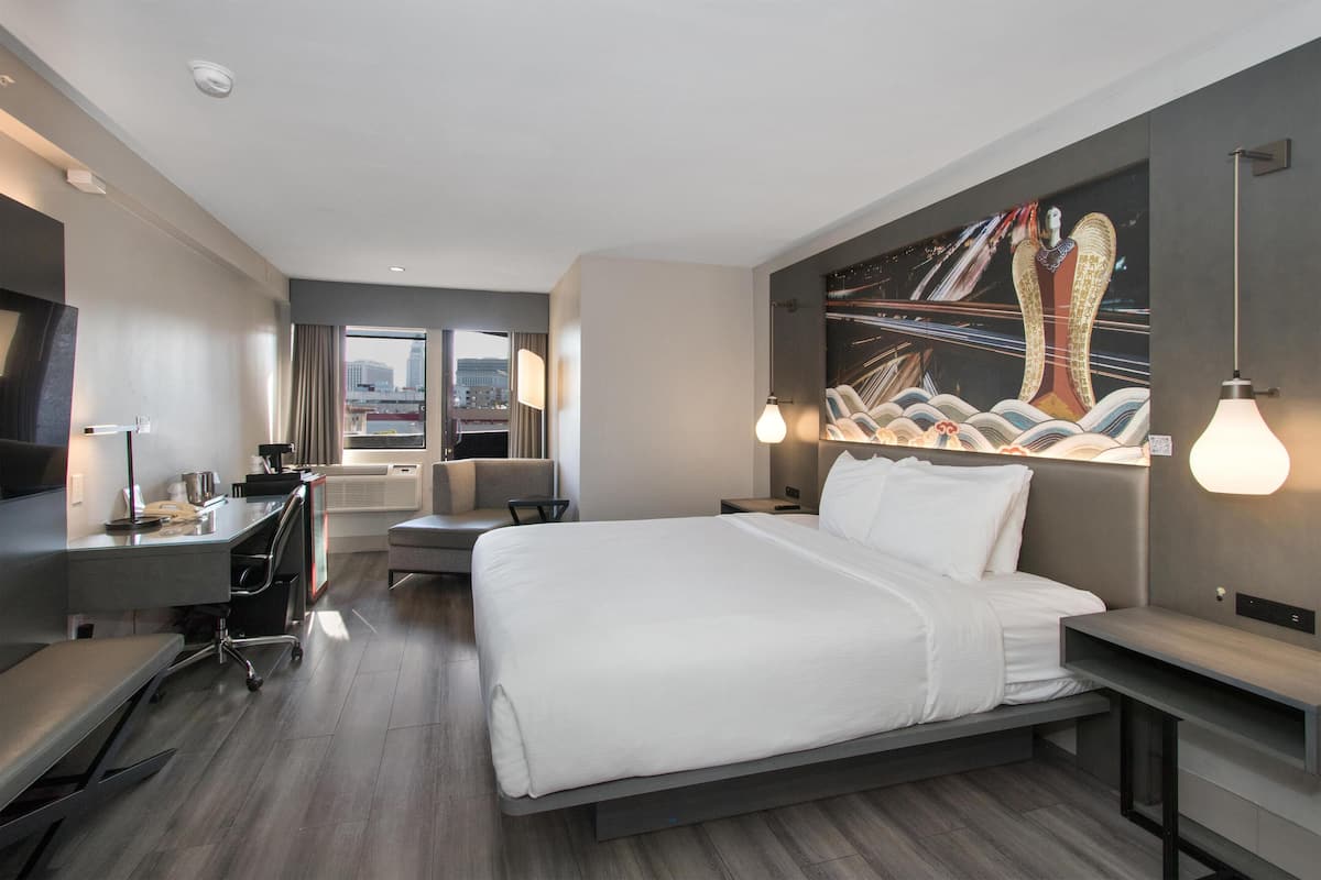 A modern hotel room with a large bed, a desk, a chair, and a TV. There is a painting on the wall and a city view through the window.