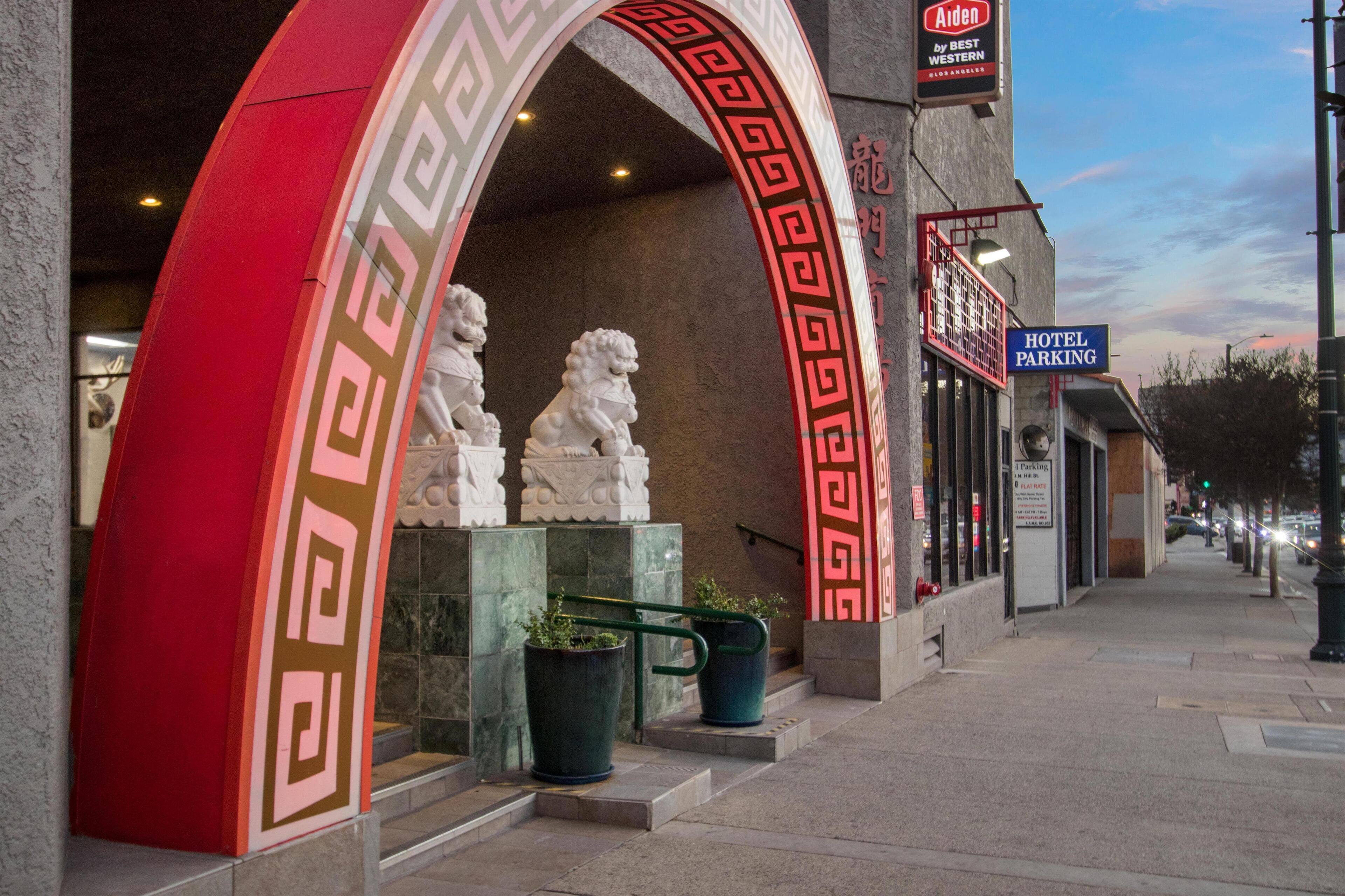L’entrée d’un hôtel, ornée d’une arche décorative rouge et blanche, avec un panneau indiquant le parking de l’hôtel et deux statues placées sur les marches.