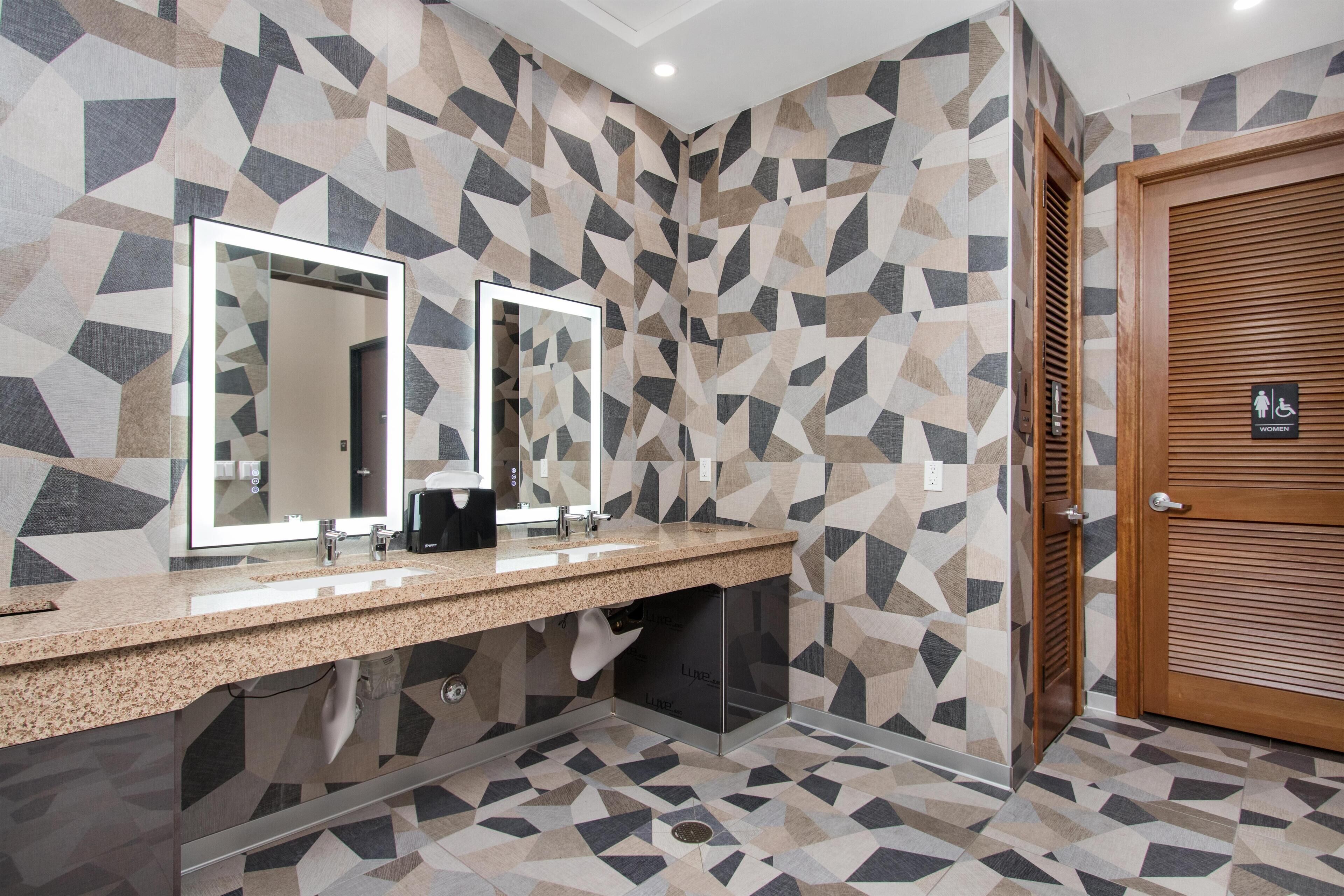 A modern bathroom with geometric patterned walls, a granite countertop, dual mirrors, and a wooden door with a women's restroom sign.