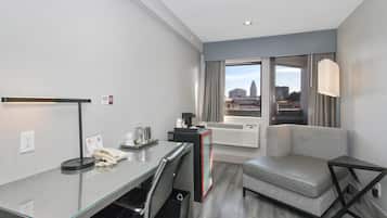 A modern hotel room with a glass desk, a sofa, a small table, and a view of the city through large windows.