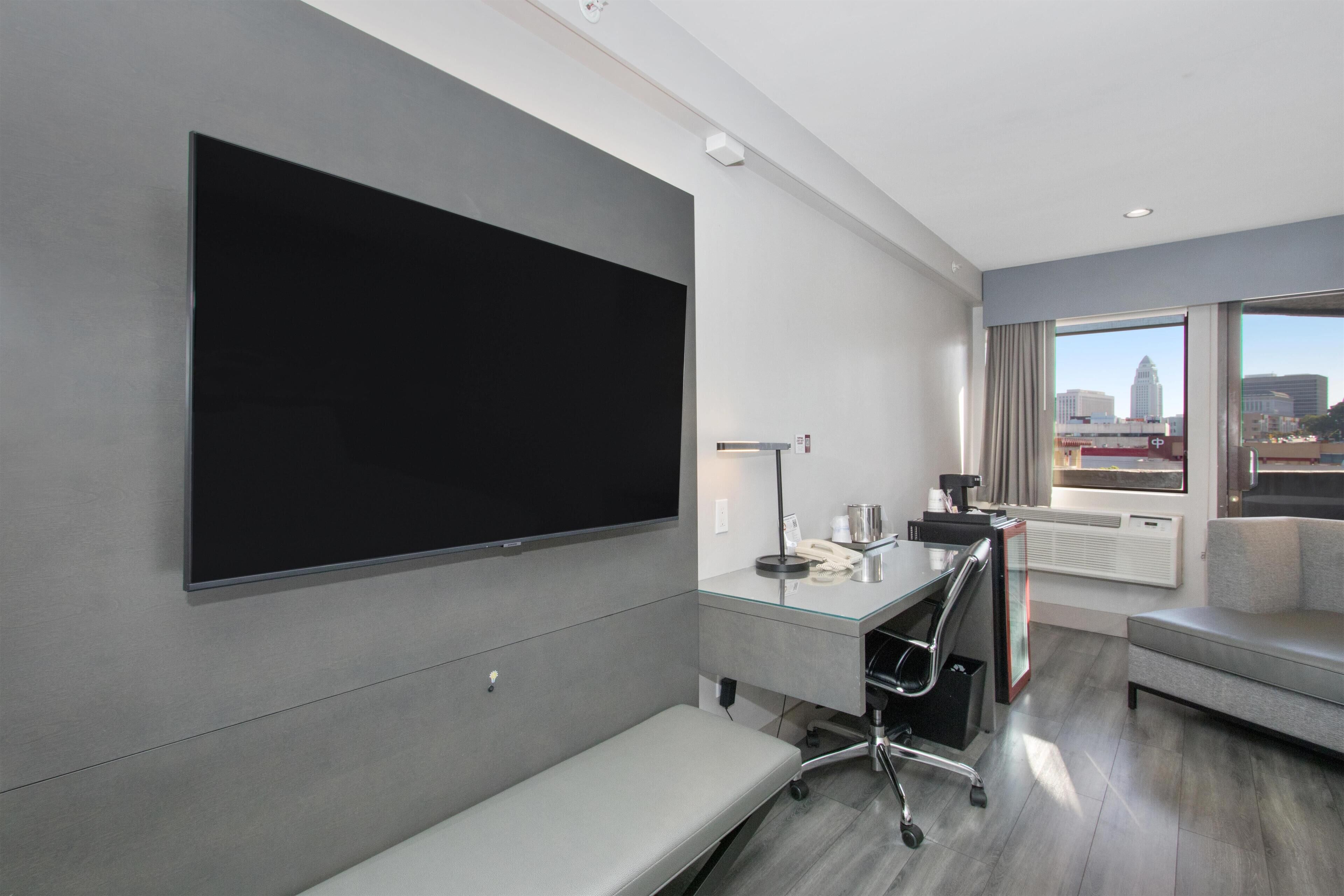 A modern hotel room with a large flat-screen TV mounted on the wall, a desk with a lamp, a chair, a sofa, and a large window offering a city view.