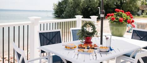 A white table set outdoors with a view of the sea, surrounded by white chairs and a pink umbrella.