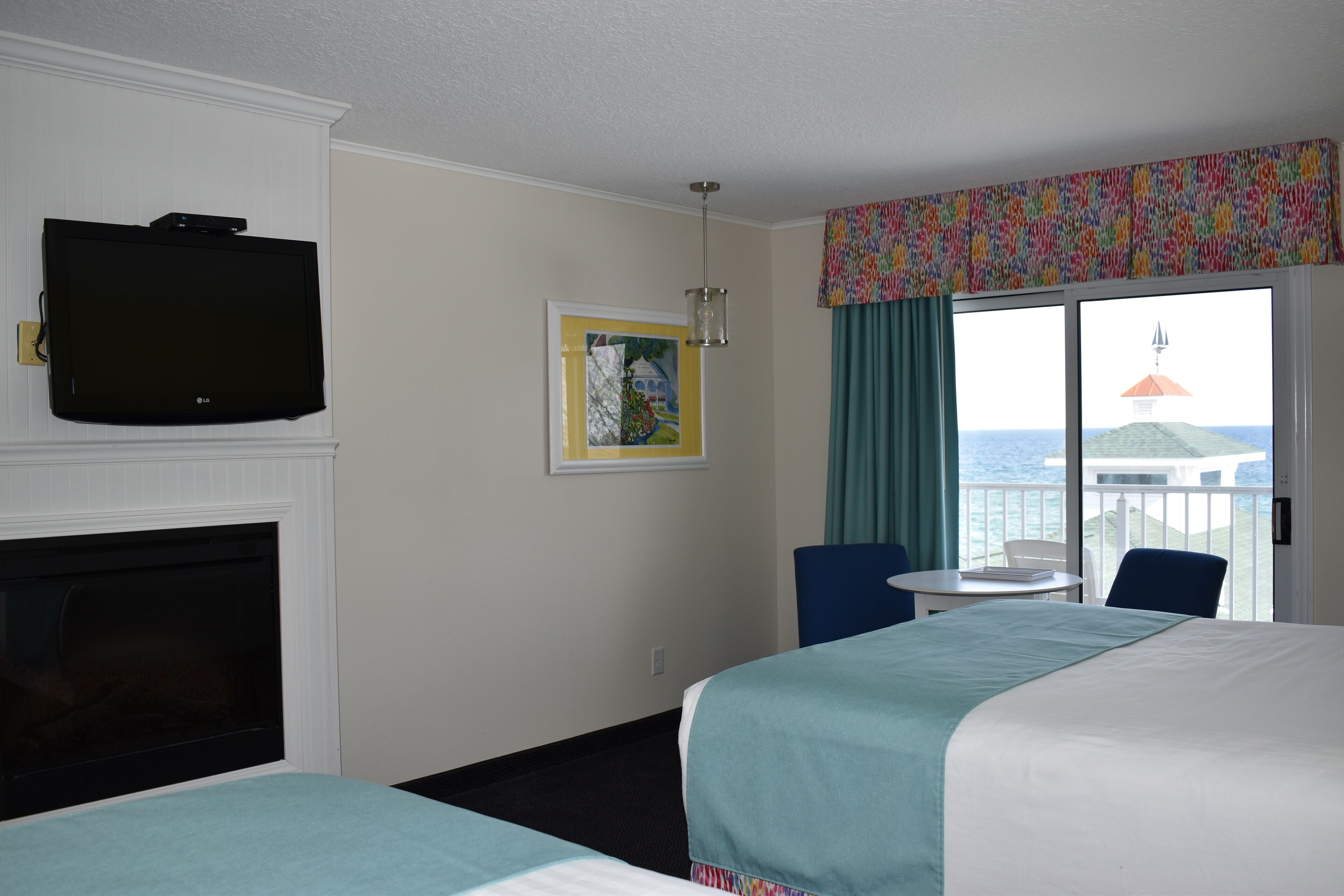 A hotel room with two beds, a TV, a fireplace, a painting, a balcony with a view of the sea, and a table with chairs.