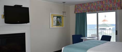 A hotel room with two beds, a TV, a fireplace, a painting, a balcony with a view of the sea, and a table with chairs.