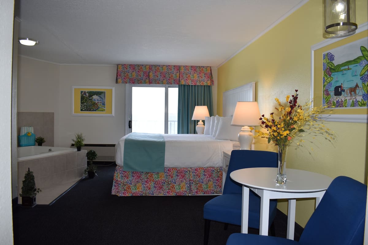 Ein Hotelzimmer mit einem Bett, einem kleinen Tisch mit einer Vase, einem Stuhl, einem Fenster mit Vorhängen, einer Wandlampe und einem Bild.