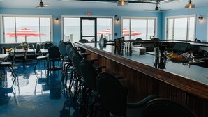 A bar area with a long counter, multiple chairs, and large windows with umbrellas. - Breakers Resort & Beach Bar (St Ignace)