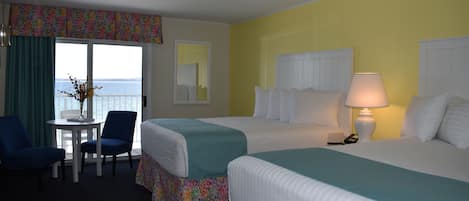 A hotel room with two beds, a small table with chairs, a lamp, and a view of the sea through a window.