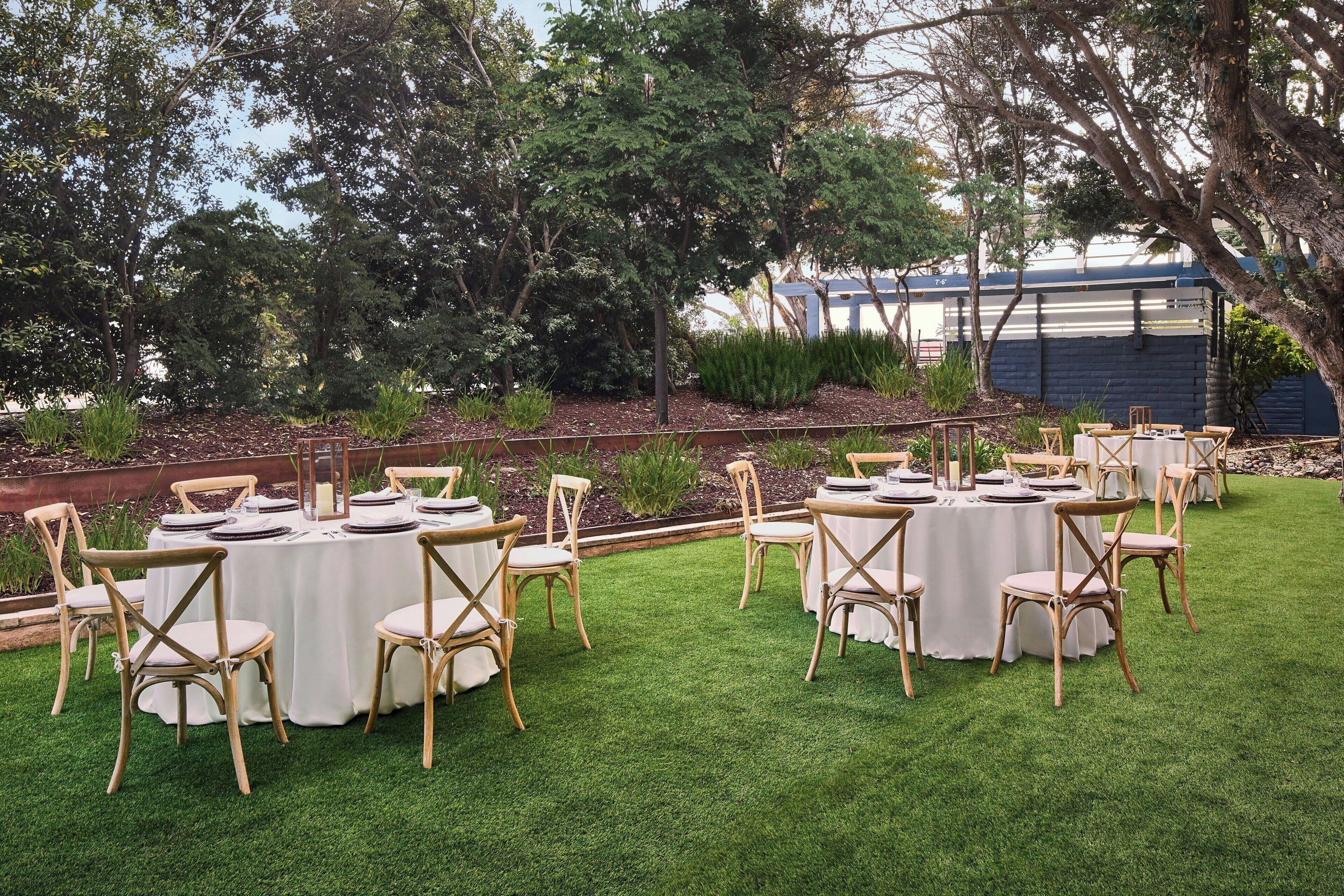 Un área de comedor al aire libre con mesas y sillas redondas, rodeada por un césped cuidado y árboles.