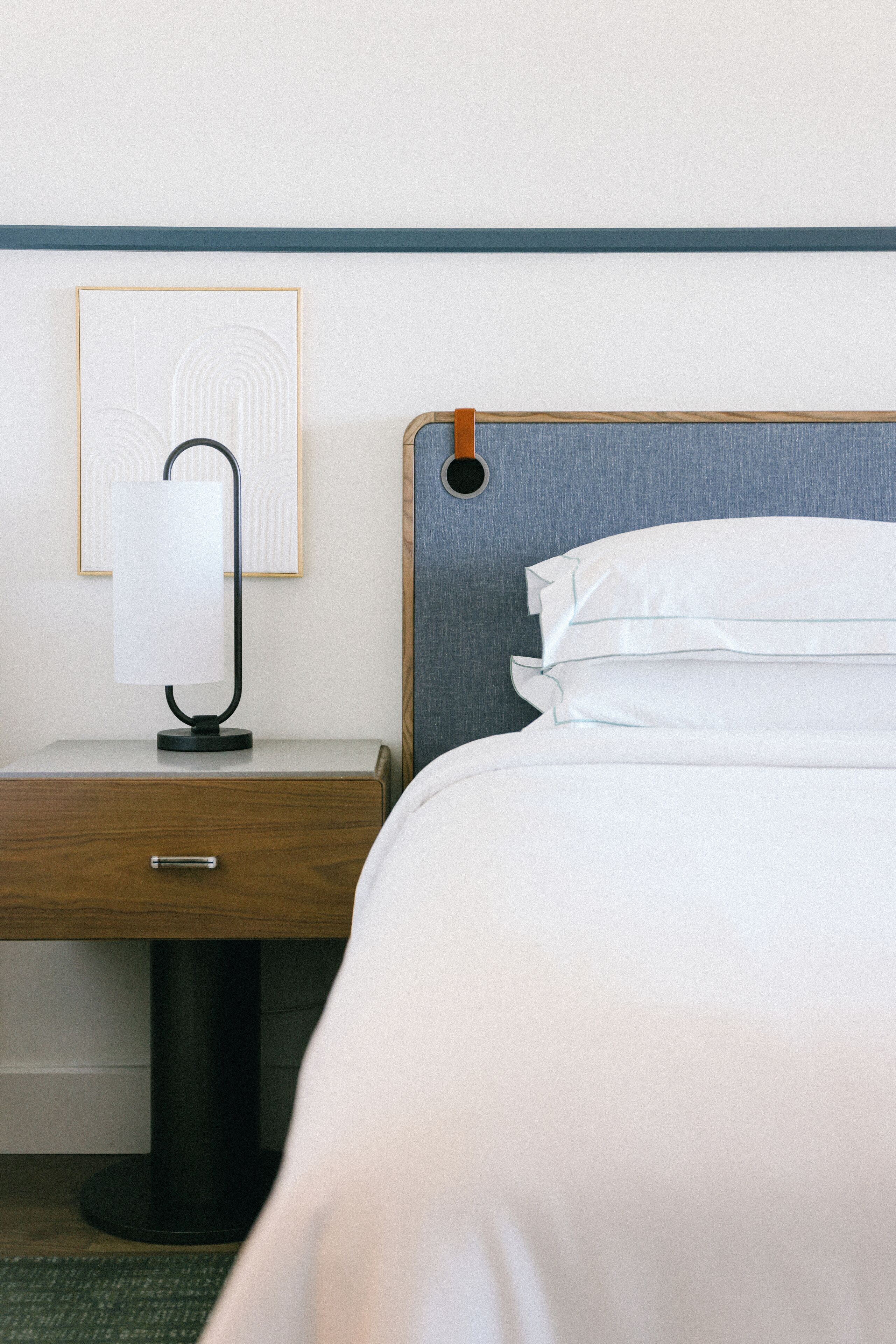 A neatly made bed with white linens, a wooden nightstand with a lamp, and a framed picture on the wall.