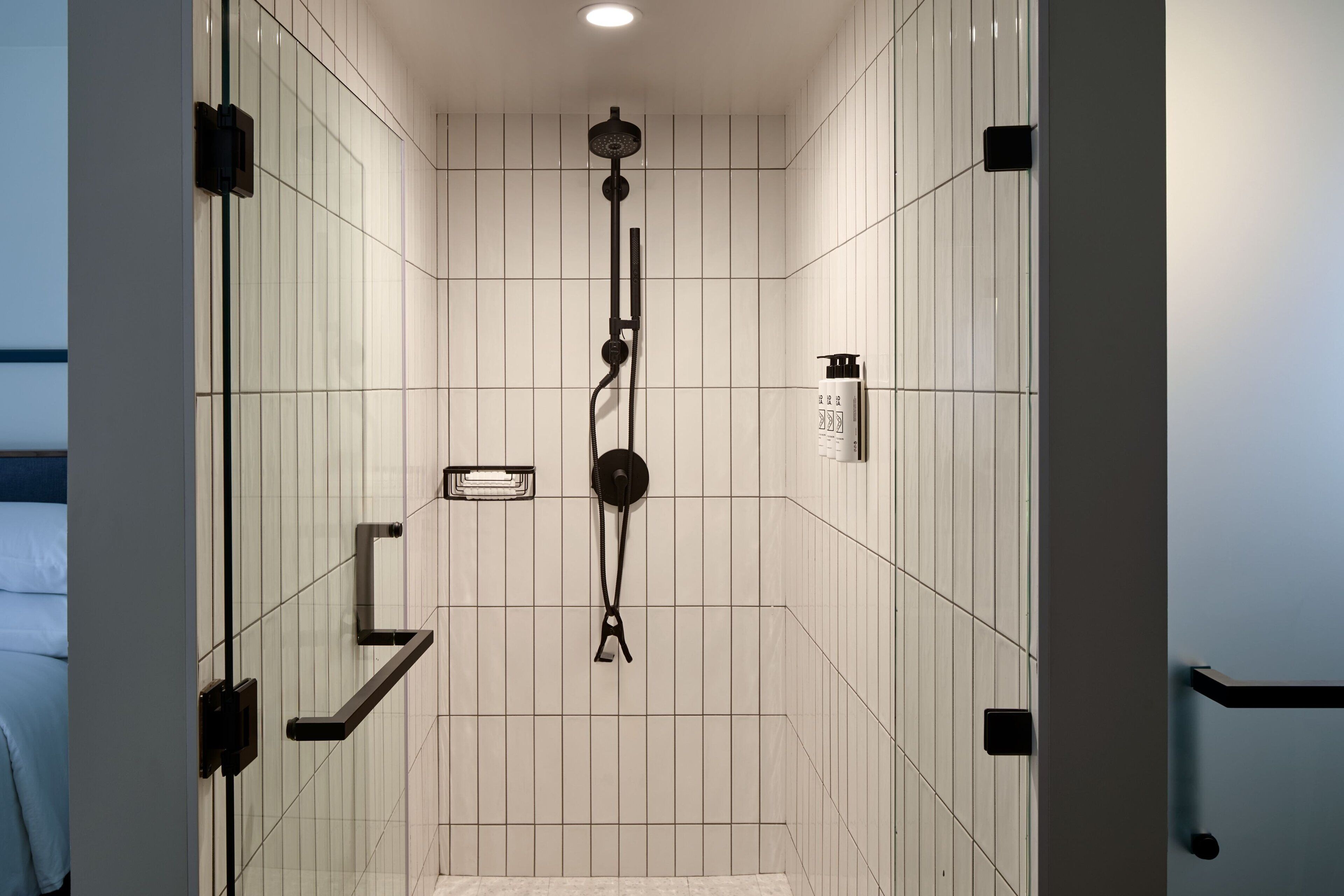 A modern bathroom with white tiled walls, a black showerhead, and a small shelf with toiletries.