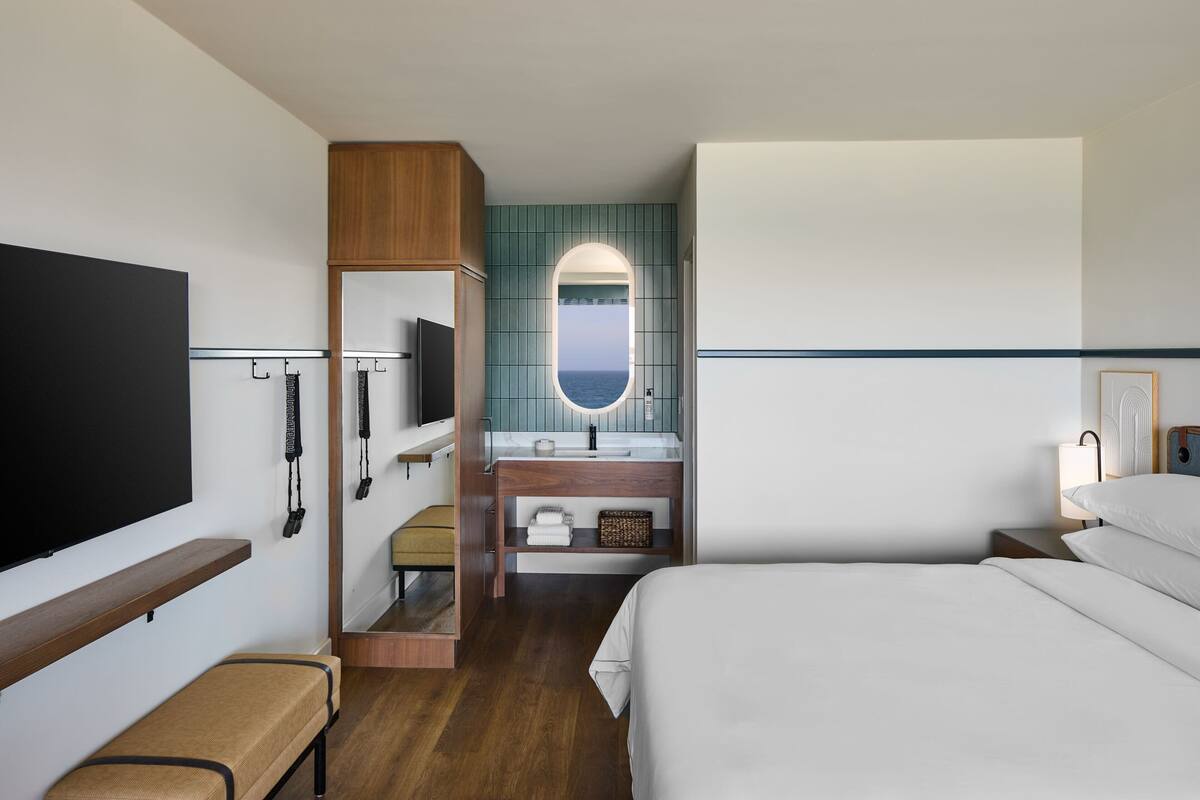 A hotel room with a bed, a TV, a bathroom with a mirror, and a view of the sea.