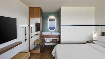 A hotel room with a bed, a TV, a bathroom with a mirror, and a view of the sea.