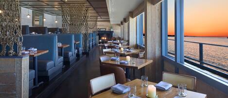 A restaurant with booths and tables, a fireplace, and a view of the sunset over the water.