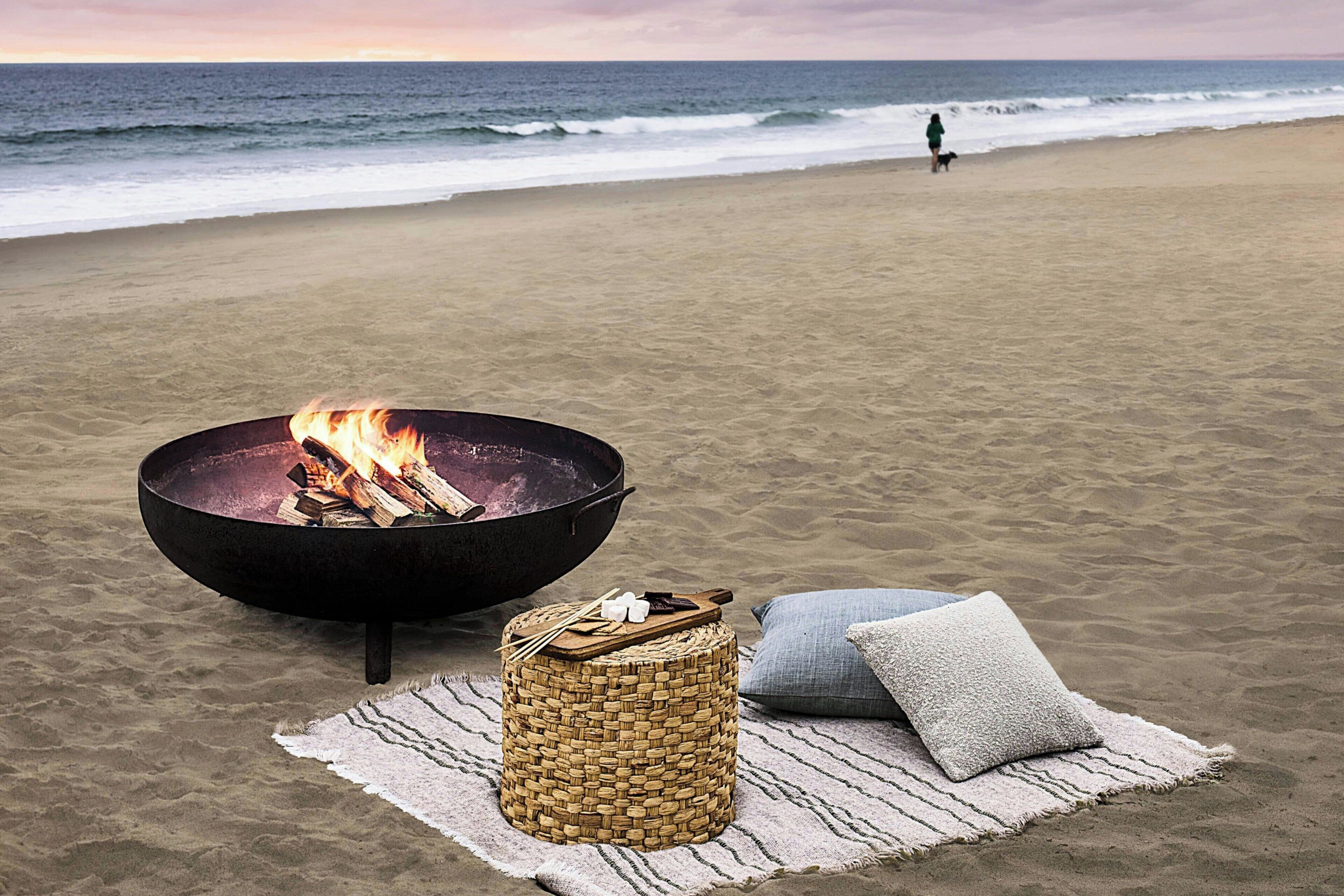 Una playa con un hogar para fuego, una cesta tejida y un cojín sobre una estera.