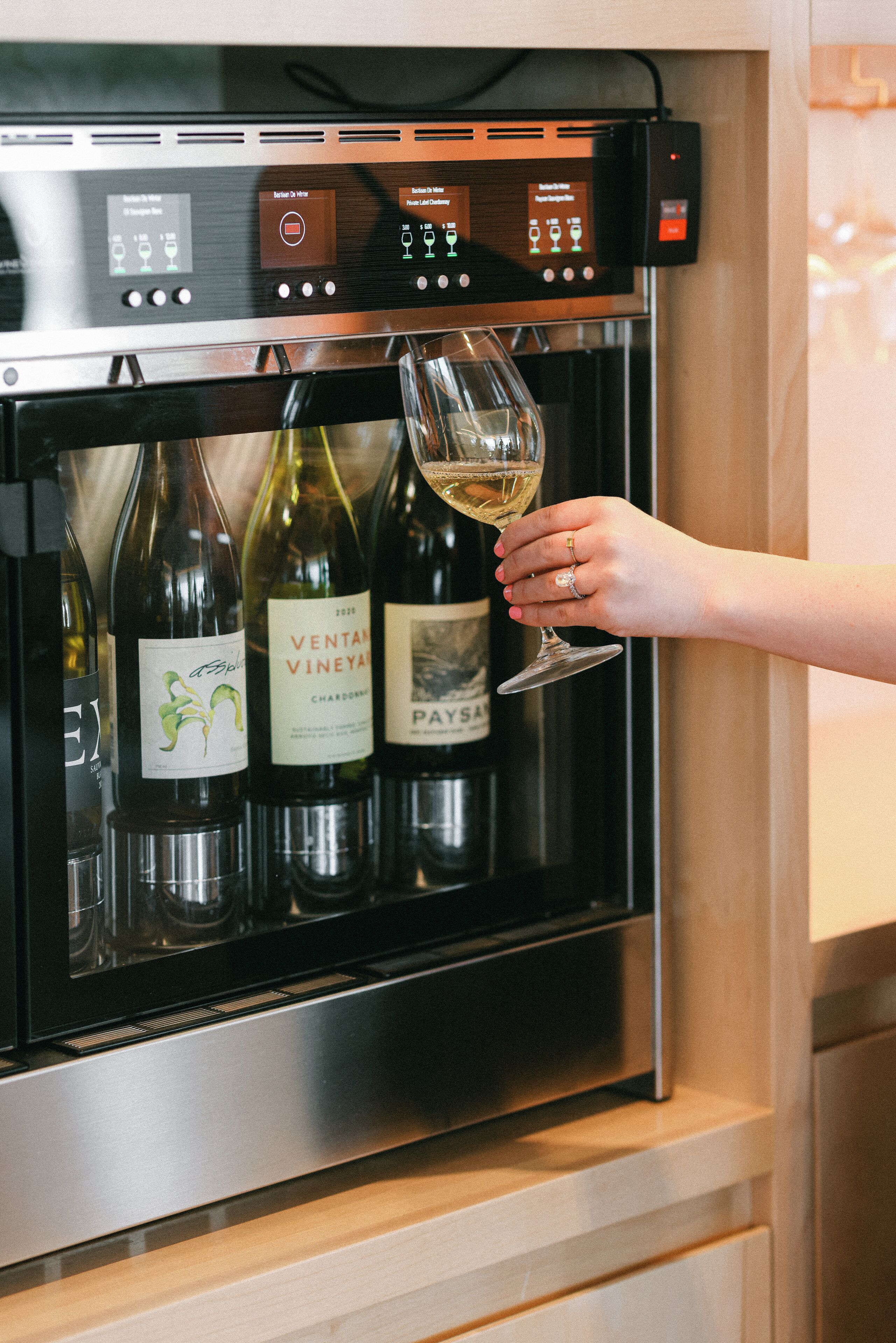 A wine dispenser with three bottles of wine and a hand holding a wine glass.