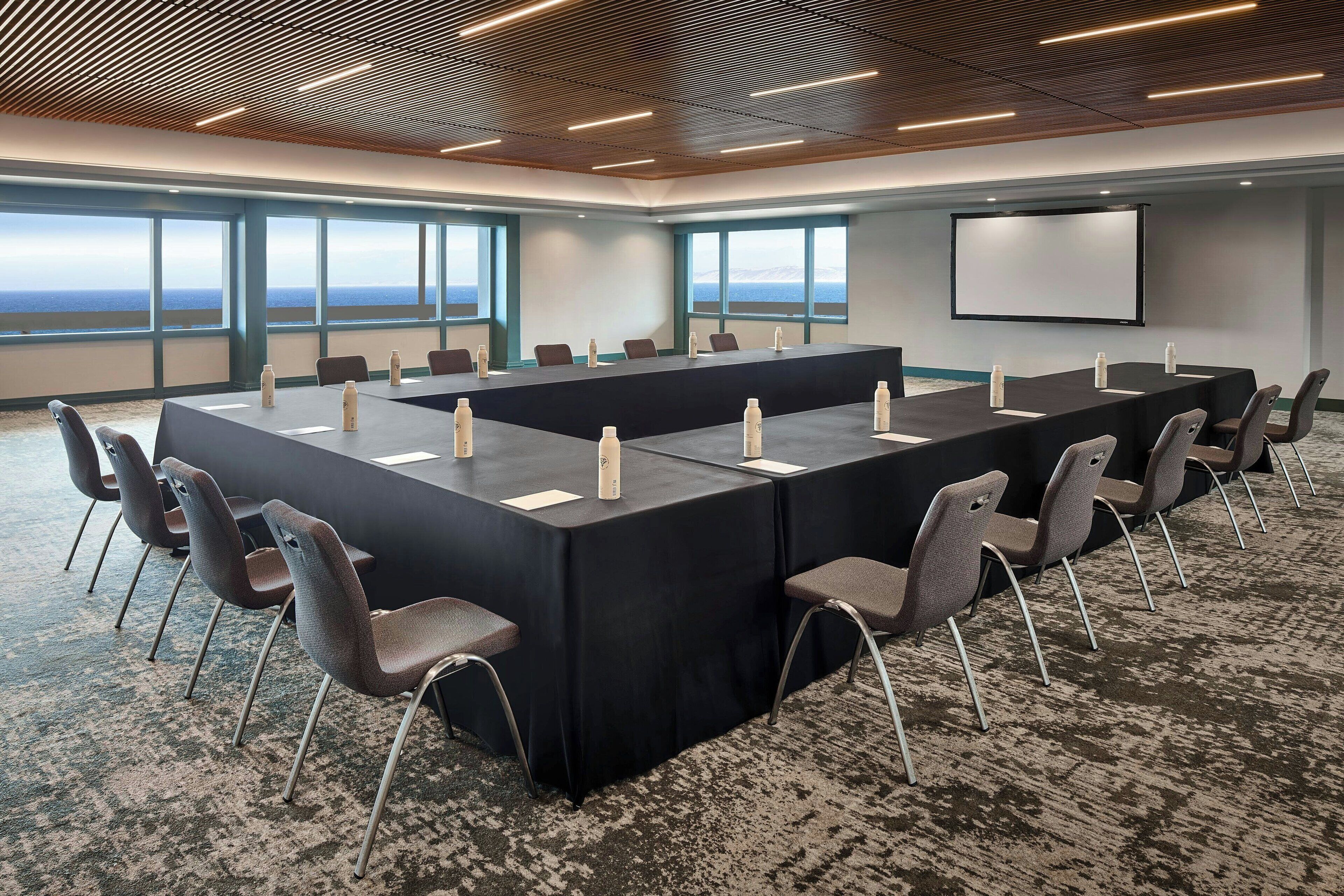 Sala de conferência com uma mesa comprida, cadeiras, tela de projetor e janelas amplas com vista.
