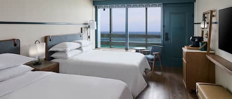 A hotel room with two beds, a desk, a chair, a TV, and a view of the ocean.