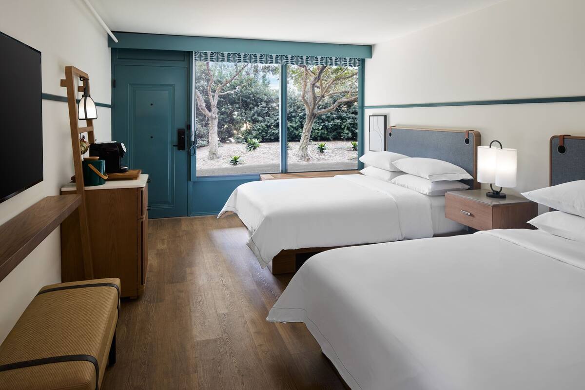 A hotel room with two beds, a desk, a chair, and a window with a view of trees.