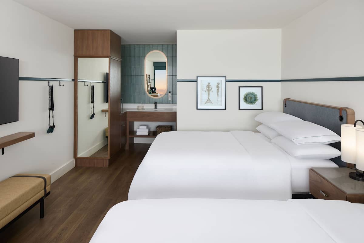 A hotel room with a bed, bedside table, lamp, and a mirror. There is a bench and a shelf with toiletries.