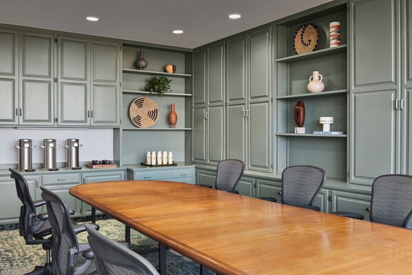 Uma sala de conferências com uma mesa de madeira comprida, seis cadeiras e armários verde-claros com prateleiras que exibem objetos decorativos.