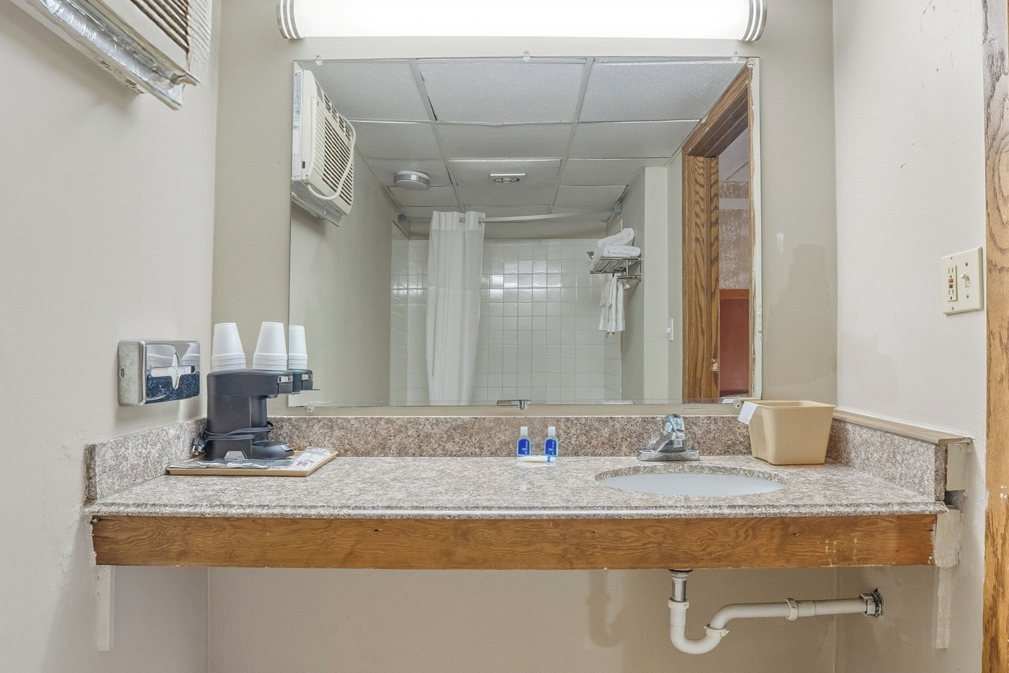 A bathroom with a granite countertop, a sink, a mirror, a coffee machine, and a shower area visible through the doorway.
