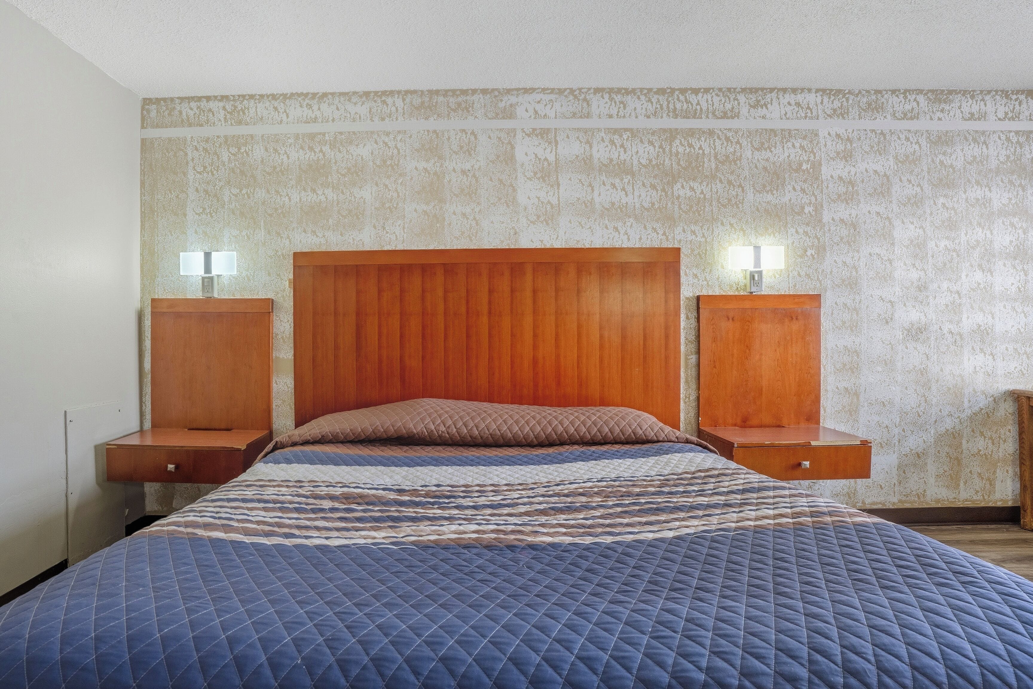 Un dormitorio con cabecera de madera, colcha azul acolchada, dos mesitas de noche con lámparas y una pared con textura.