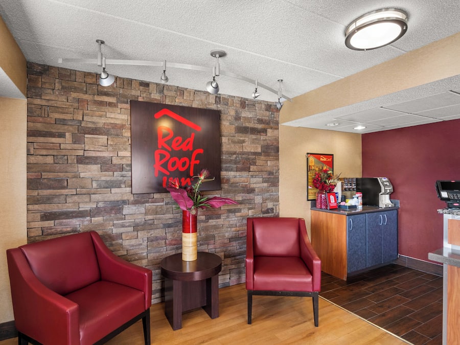 A Red Roof lounge area with two red chairs, a small table with a vase, a brick wall, a reception desk, and a coffee machine.