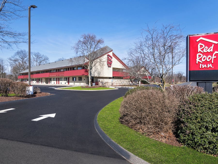 Un Red Roof Inn con fachada roja y blanca, un estacionamiento con flechas direccionales y un cielo azul despejado.