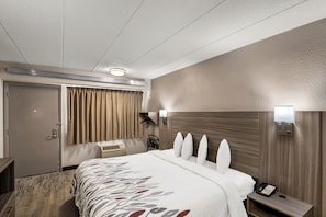 Une chambre d’hôtel avec un grand lit, une table de chevet, une lampe, un téléphone fixé au mur et une fenêtre avec des rideaux.