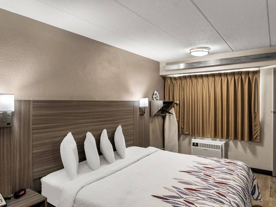 Ett hotellrum med en säng, ett nattduksbord med en klocka, en garderob, en spegel och ett fönster med gardiner.