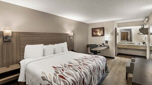 A hotel room with a large bed, bedside tables, a desk, a chair, a desk lamp, a wall-mounted TV, and a bathroom with a sink and mirror. - Red Roof Inn Enfield (Enfield)