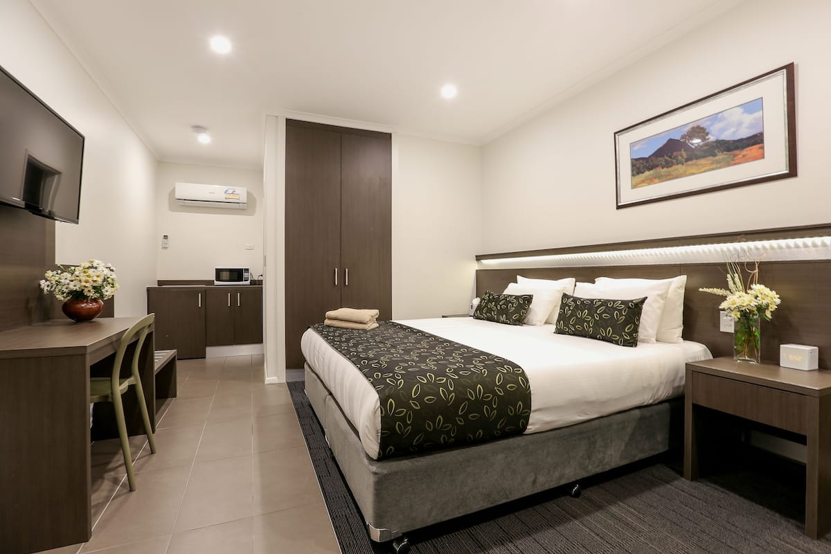 A modern hotel room with a bed, bedside table, and a desk with a chair. There is a microwave and a cabinet in the corner.