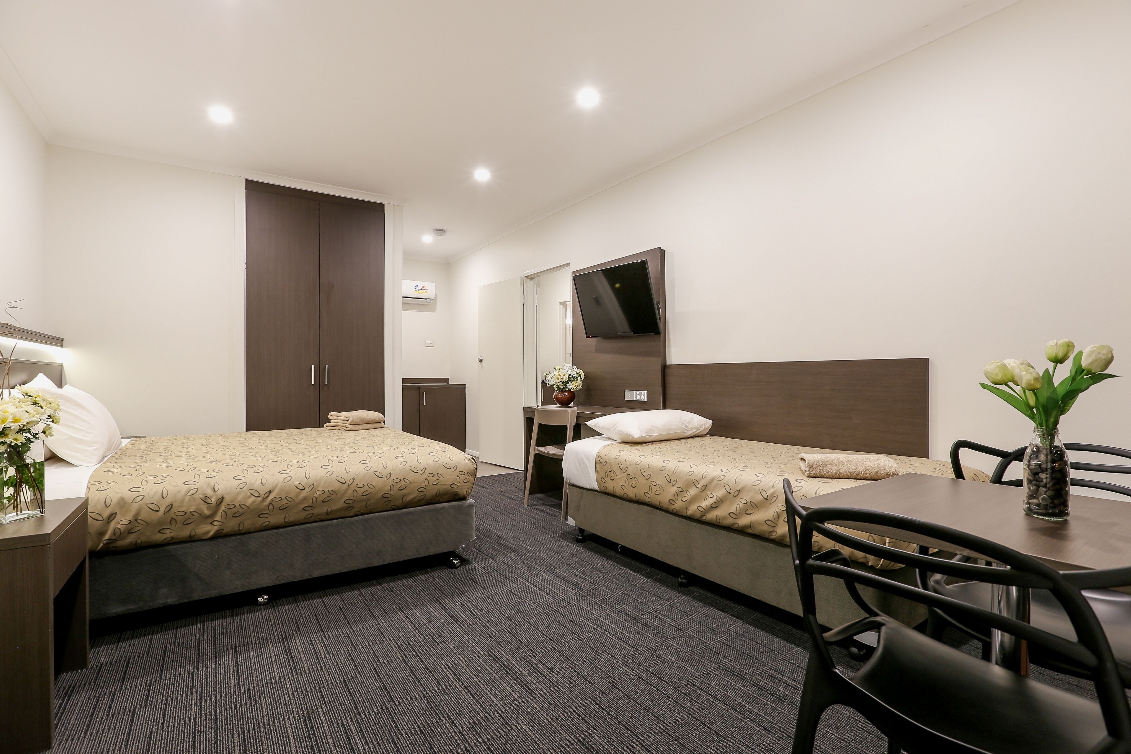 A hotel room with two beds, a desk, a chair, a TV, and a vase with flowers.