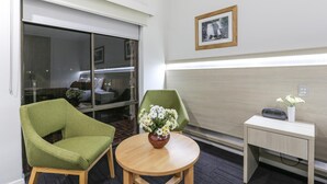 A hotel room with a round wooden table, two green armchairs, a small white side table with a vase of flowers, and a framed picture on the wall. - Morphettville Motor Inn (Camden Park)