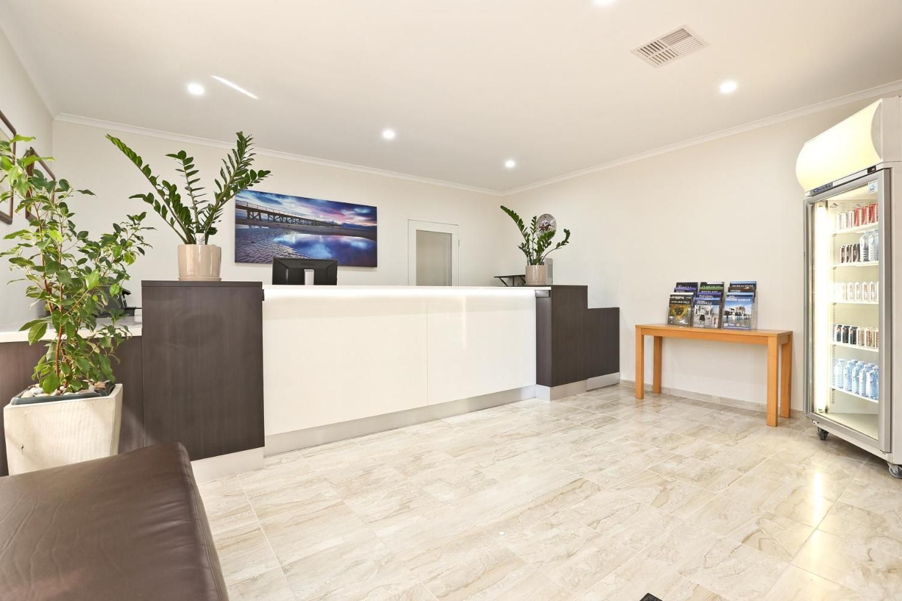 A reception area with a white reception desk, potted plants, a wall-mounted TV, and a refrigerator stocked with beverages.