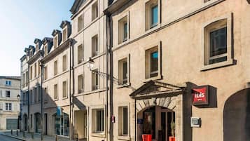 La façade d’un hôtel présente un style architectural classique, avec de nombreuses fenêtres et une entrée imposante surmontée d’une enseigne rouge.