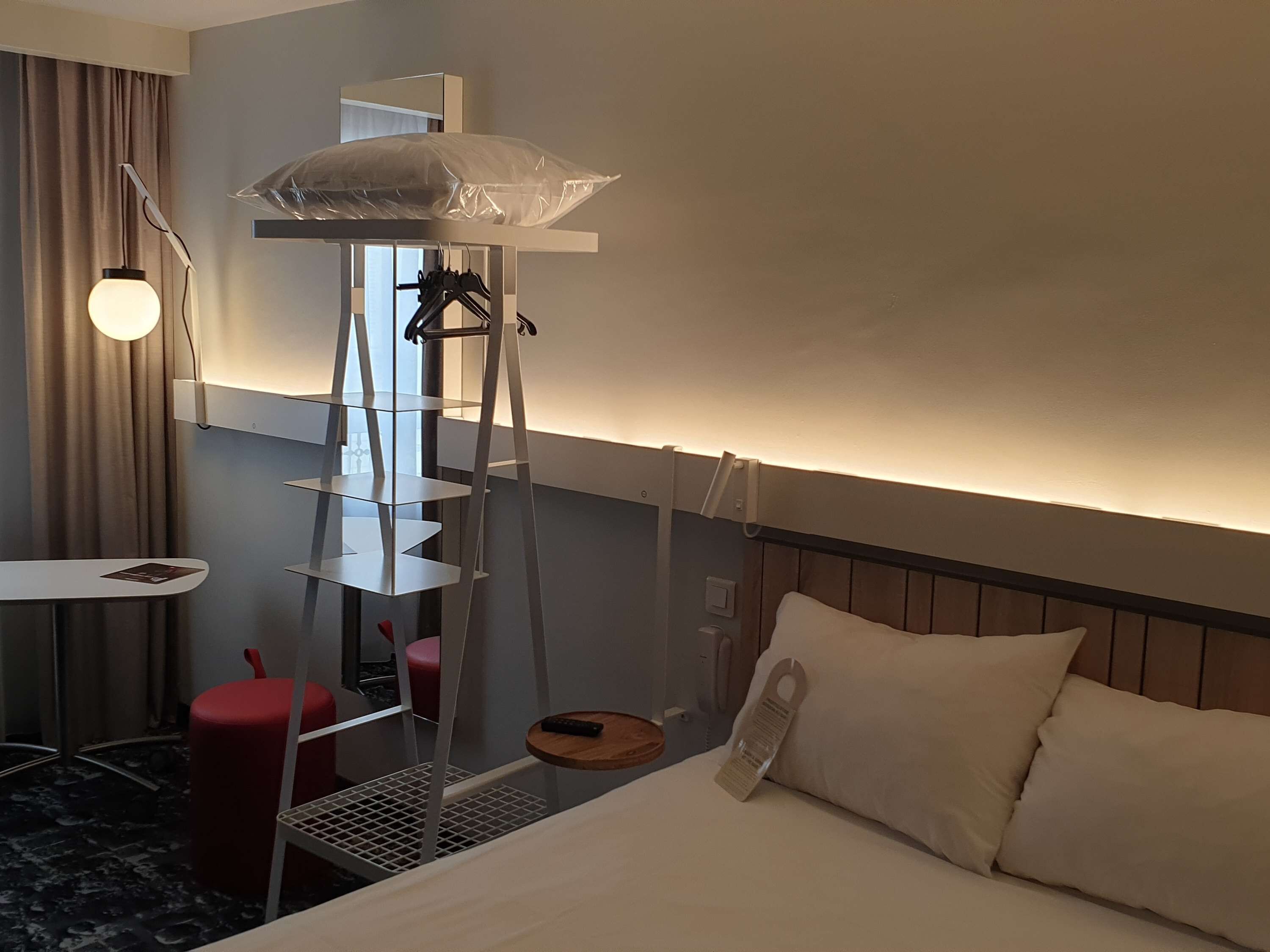 A hotel room with a bed, a small table, a red chair, a white shelf unit, and a hanging light fixture.