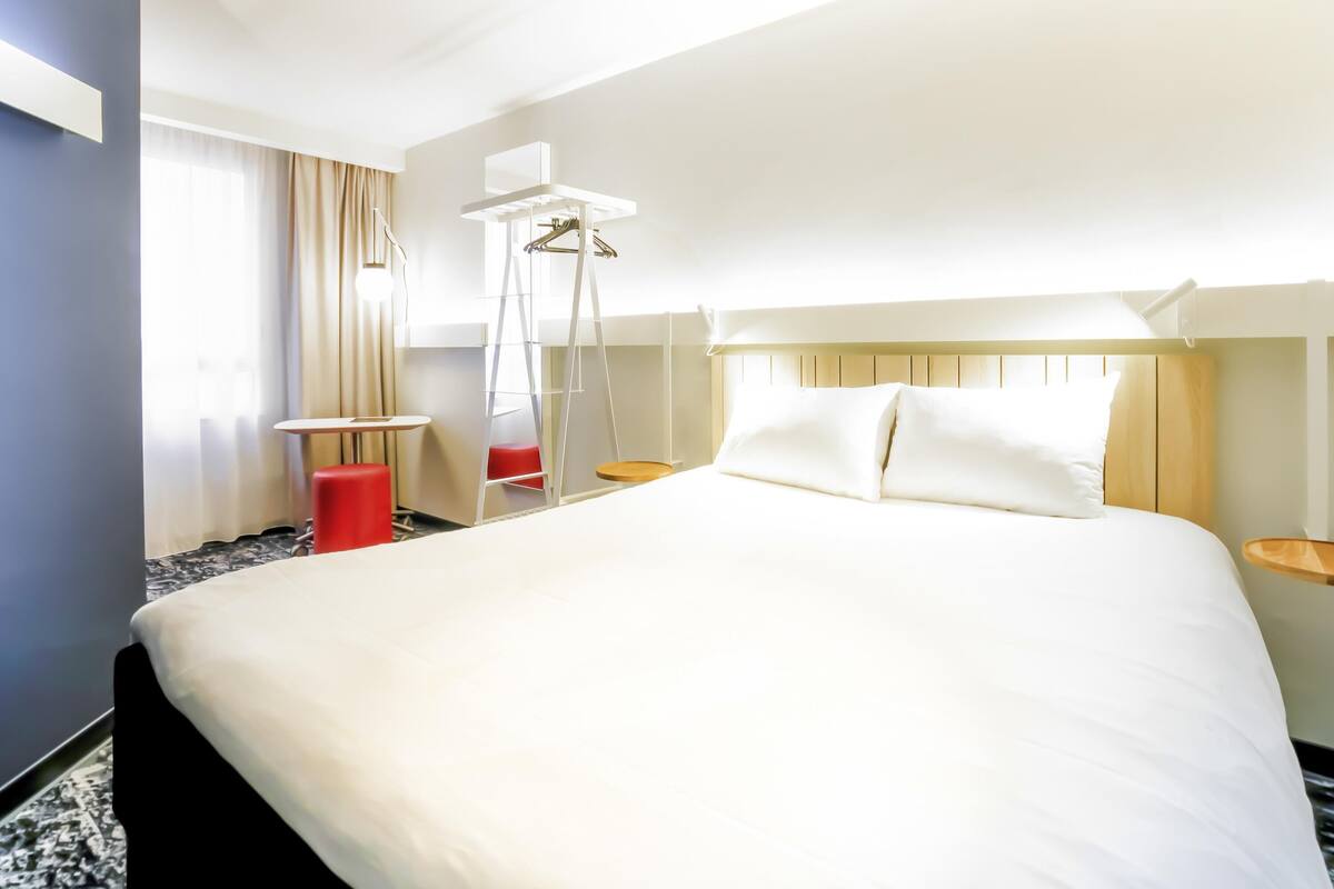 A hotel room with a large bed, a small table, a chair, a hanging light fixture, and a window with curtains.