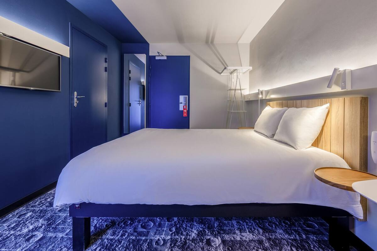 A modern hotel room with a large bed, a flat-screen TV mounted on the wall, a bedside table with a lamp, and a wooden headboard.