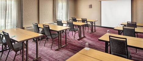 A conference room with multiple tables and chairs, a projector screen, and large windows with curtains.