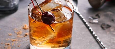 Un vaso de bebida color ámbar con cubitos de hielo y una cereza en brocheta, sobre una superficie de madera con un coctelero y una botella al fondo.