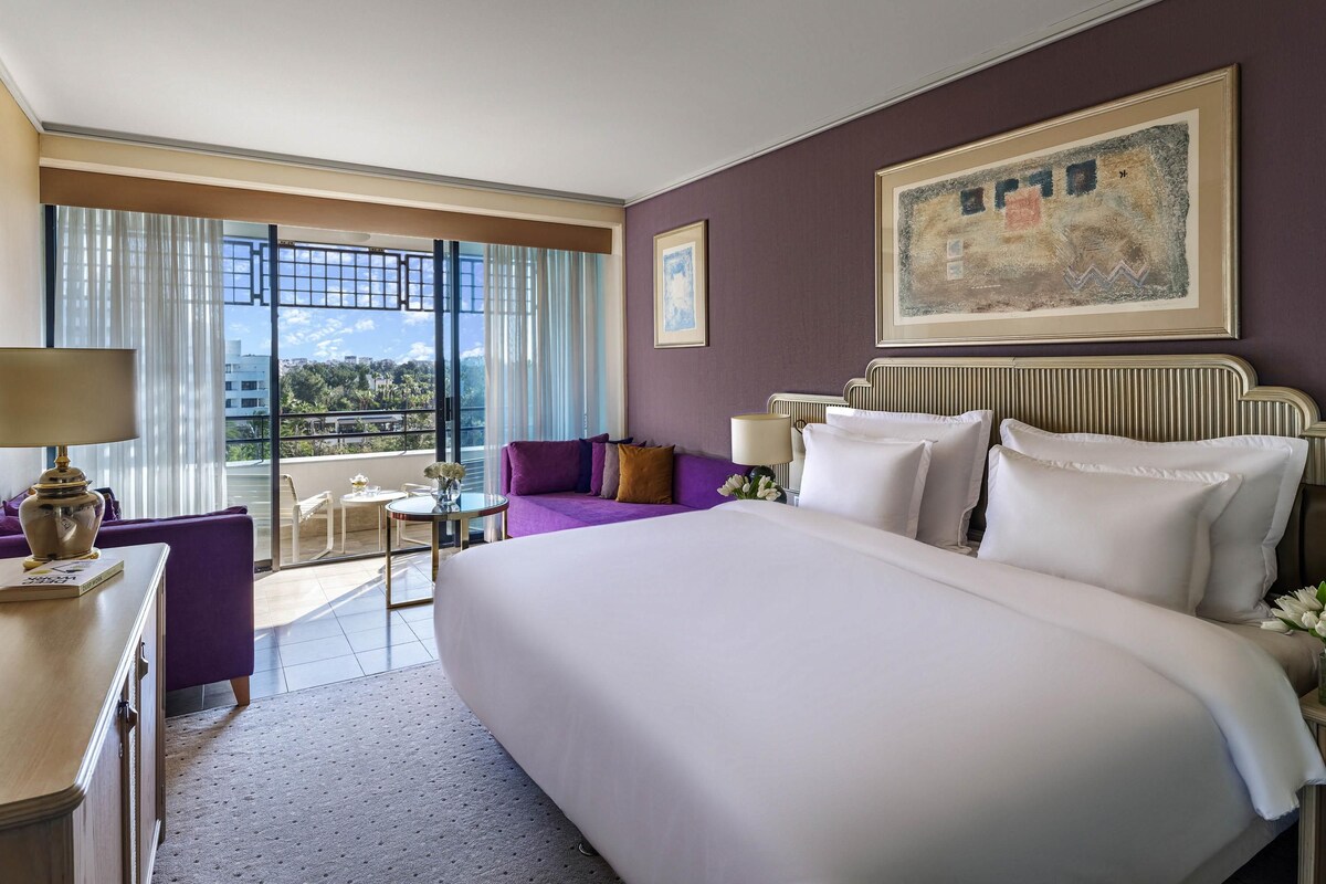 A hotel room with a large bed, a purple sofa, a small table with a lamp, and a view of the city through large windows.
