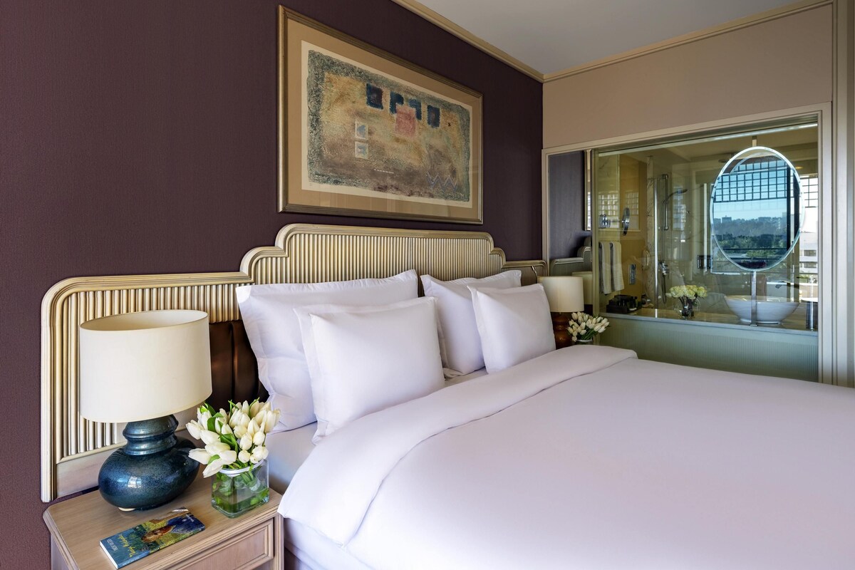 A hotel room with a large bed, a nightstand with a lamp and flowers, a framed picture on the wall, and a bathroom with a mirror and shower.