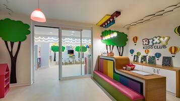 Een kinderclub met een receptiebalie, kleurrijk zitmeubilair en speelse wanddecoraties, waaronder bomen en luchtballonnen.
